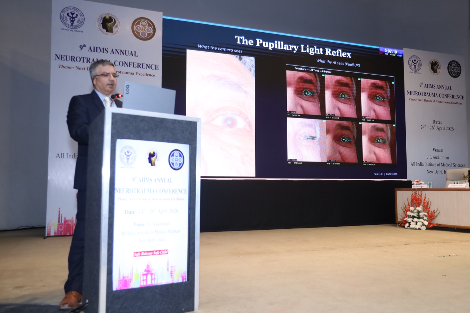 Dr. Ajay Bakshi presenting PupiLUX at the 9th AIIMS Annual Neurotrauma Conference, with the Pupillary Light Reflex slide projected on stage.